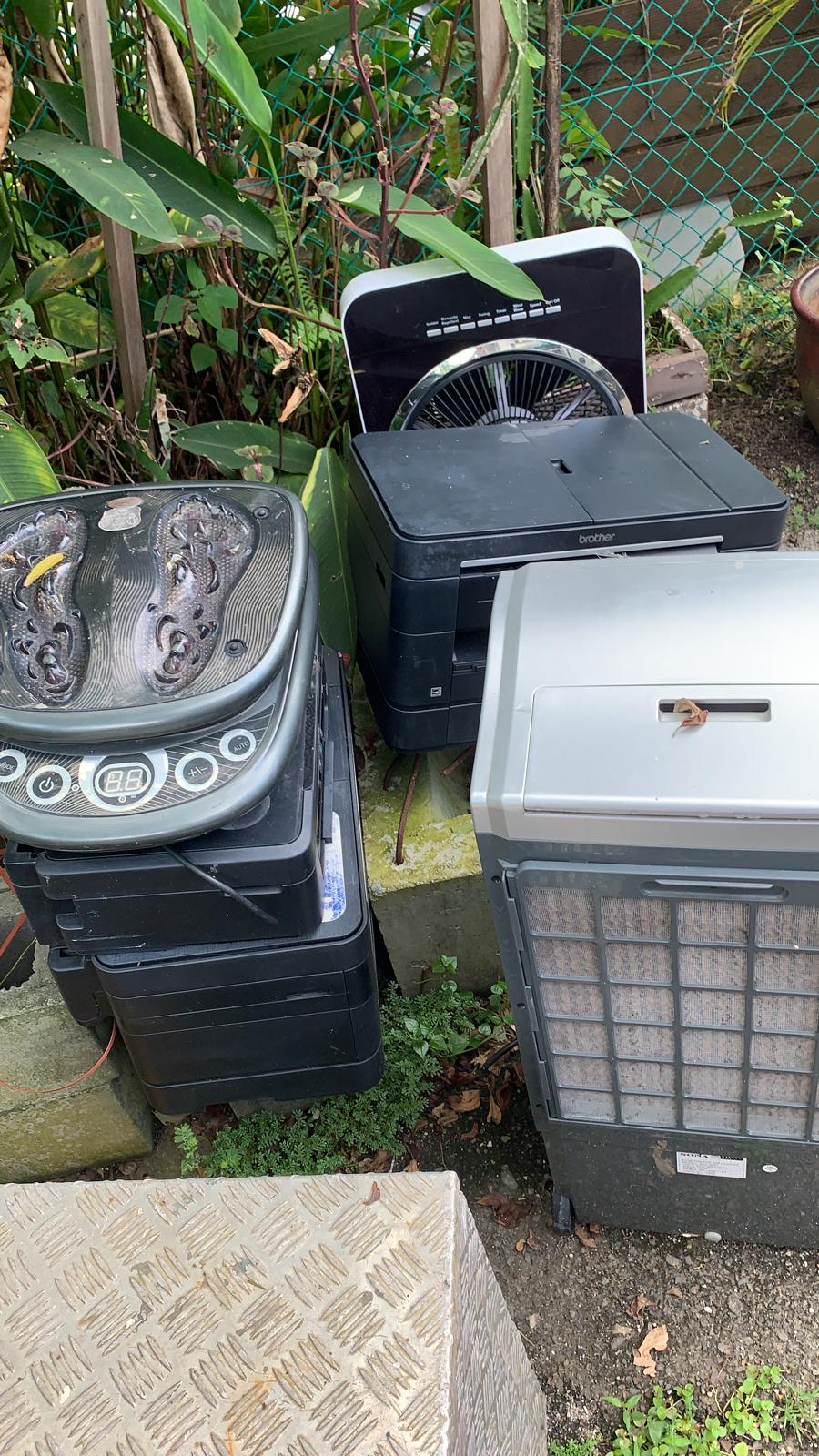 Pile of discarded electronics including printers, a desk fan, and a foot massager stacked outdoors.